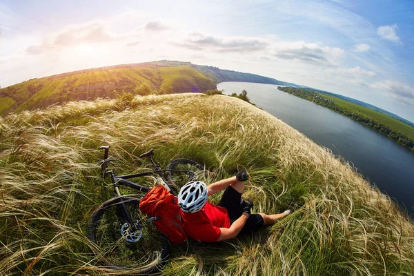 Junger Radfahrer rastet mit Mountainbike gegen schönen Himmel auf der Wiese aus. — Stockfoto