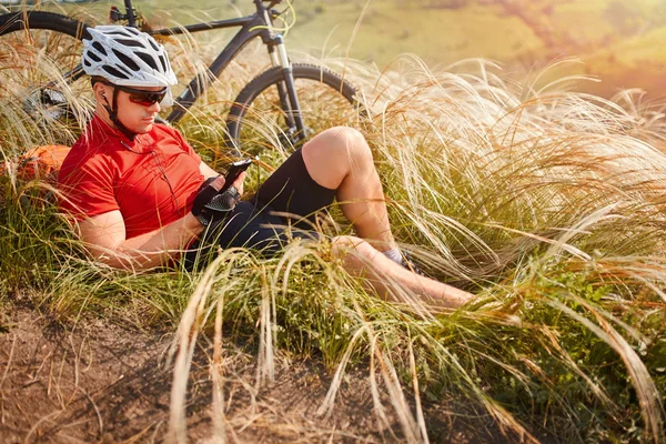 Biker in red t-shirt white helmet with backpack resting under a tree with his bike