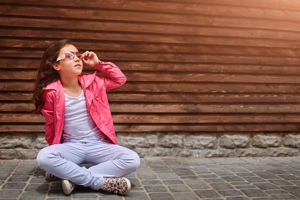 Κομψό μικρό παιδί κορίτσι φορώντας ένα ροζ σακάκι, καλοκαίρι ή φθινόπωρο, λευκό τζιν, γυαλιά ηλίου — Φωτογραφία Αρχείου