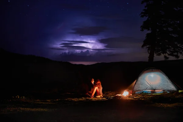 Night camping in the mountains. Couple tourists have a rest at a campfire near illuminated tent