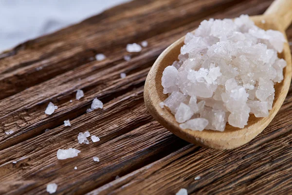 Crystal sea salt in a wooden spoon on dark vintage wooden background, top view, close-up, selective focus.