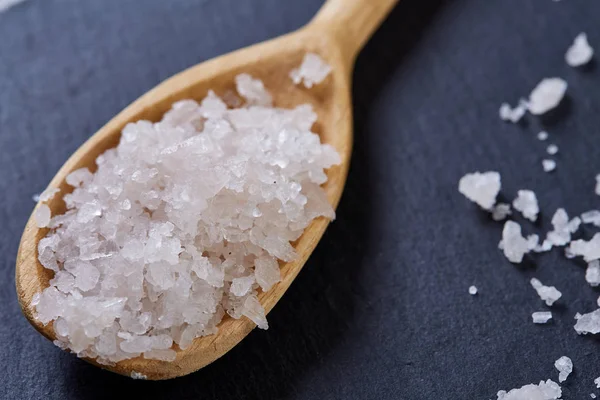 Crystal sea salt in a wooden spoon on dark background, top view, close-up, selective focus.