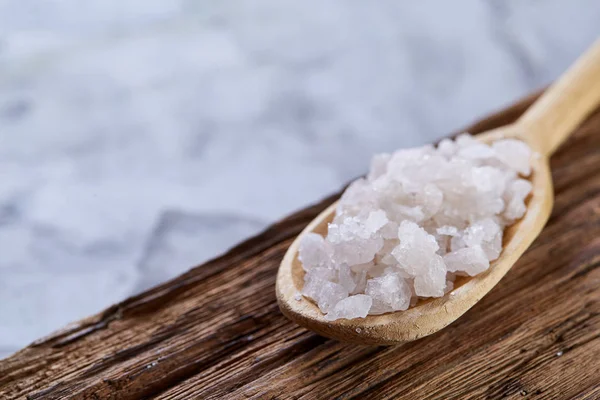 Crystal sea salt in a wooden spoon on dark background, top view, close-up, selective focus.