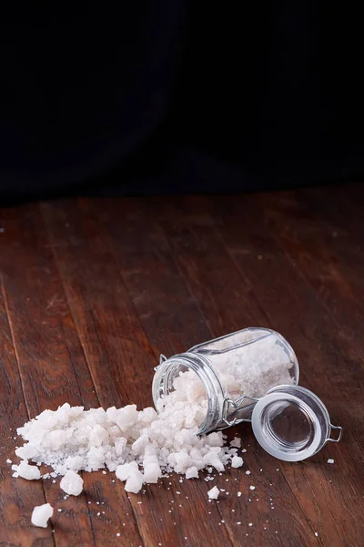 Spa concept. Bath salt pouring out of glass jar on wooden table, close-up, selective focus — Stock Photo, Image