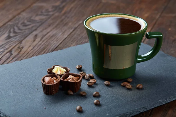 Taza de café, granos de café, caramelos de chocolate en tablero de piedra sobre fondo de madera, enfoque selectivo, primer plano —  Fotos de Stock