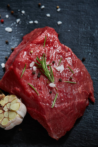 Raw meat beef steaks on black slate board with spices, garlic and rosemary over wooden background, selective focus