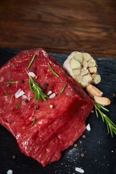 Rauw vlees rundvlees biefstuk op zwarte leisteen bord met specerijen, knoflook en rozemarijn op houten achtergrond, selectieve aandacht — Stockfoto