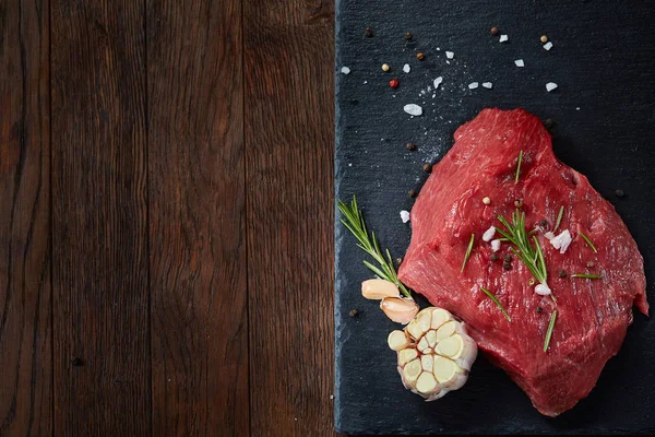 Rauw vlees rundvlees biefstuk op zwarte leisteen bord met specerijen, knoflook en rozemarijn op houten achtergrond, selectieve aandacht — Stockfoto