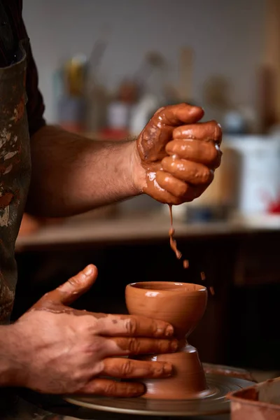 Крупным планом руки гончара в фартуке проливают немного воды, делая вазу из глины, избирательный фокус — стоковое фото