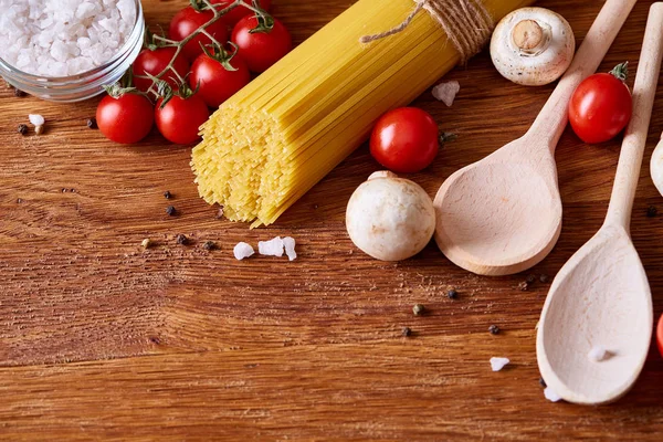 Pâtes alimentaires non cuites, tomates et deux cuillères sur fond bois, vue de dessus, gros plan, mise au point sélective — Photo