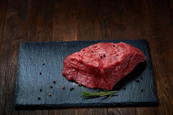 Steki z wołowiny surowego mięsa na czarny Łupek deska z przyprawami i rozmarynem na podłoże drewniane, miejsce — Zdjęcie stockowe