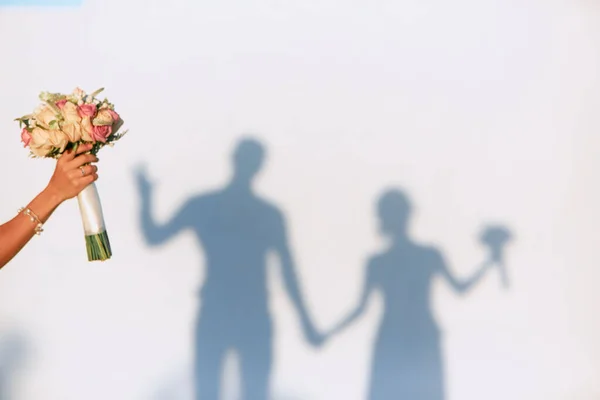 Bilezik takmış bir el düğün buketi tutuyor. Beyaz arka planda el ele tutuşan bir kadın ve erkeğin gölgeleri..