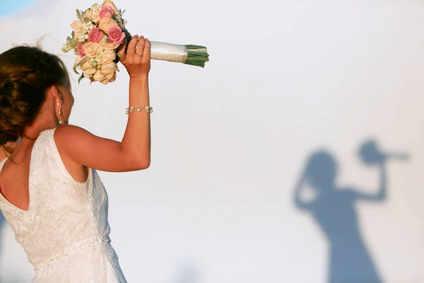 Elinde buketi olan siyah saçlı bir gelin gölgesi olan beyaz bir duvarın önünde duruyor..