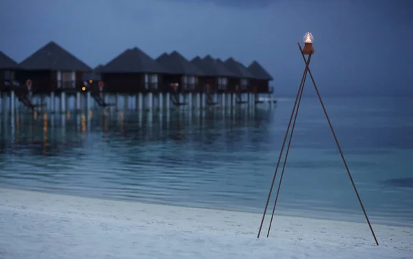 Tripodun üzerinde yanan bir mum, kumlu bir plajda, deniz ve su bungalovlarının arka planında duruyor.