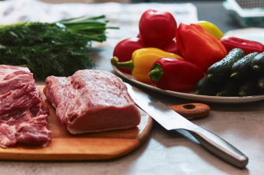 Kesme tahtasının üzerinde birkaç parça et var. Tahtanın yanında salatalık, domates, biber, maydanoz ve soğanlı tabaklar var.,