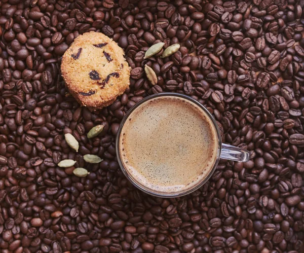 Kavrulmuş kahve çekirdekleri, köpüklü kahve kupası, kurabiyeler ve kakule taneleri.