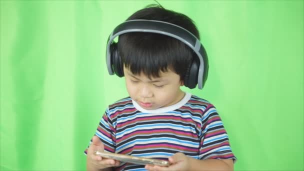 スタジオで立っているかわいいアジアの少年彼スマート フォン イヤホンで音楽を聞いている間. — ストック動画