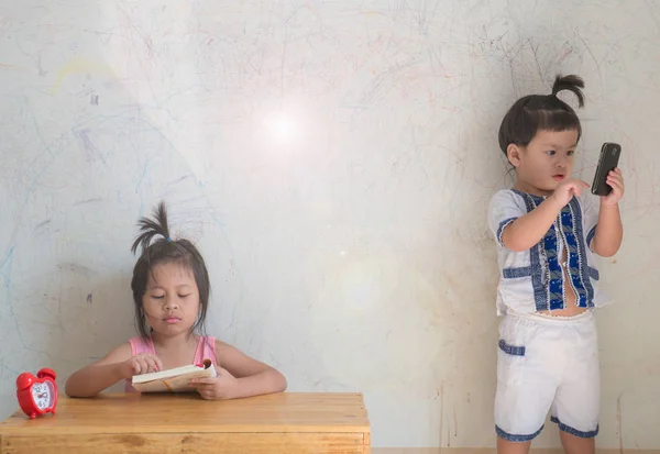 Menina leitura livro e menino jogar smartphone, parede suja fundo — Fotografia de Stock