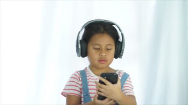 Şirin bir kız şirin görünüyor ve desenli bir gömlek giyiyor ve siyah bir örgü takıyor kulaklık takıyor, akıllı telefondan sosyal medyadan müzik dinliyor.,
