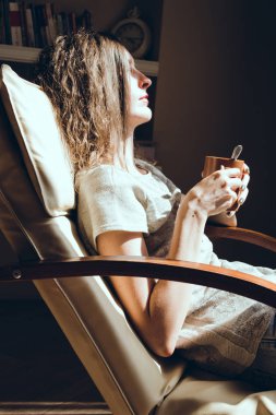 Evde zaman zevk. Kadın portre rahatlatıcı pencere holding bir fincan çay veya kahve yakın konforlu modern sandalyede kapatın. Doğal ışık. Rahat ev. Anın tadını çıkar. Parlak güneşli bir günde. Fincan sıcak içecek