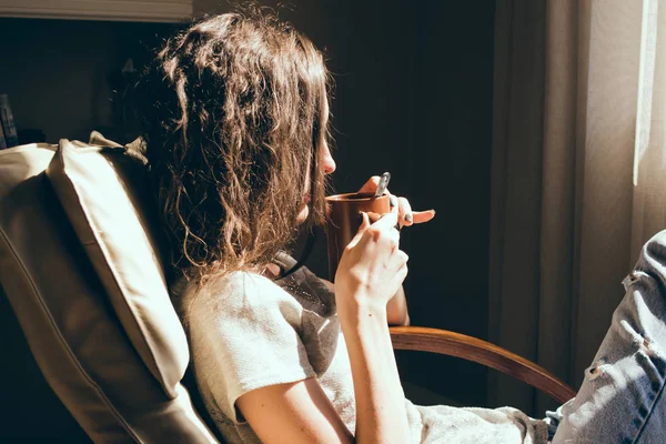 Kadın portre rahatlatıcı çay içme oturma odası penceresinde yakın konforlu modern sandalyede kapatın. Sıcak doğal ışık. Rahat ev. Casual giyim. Oda iç. Düşünceli kadın. Parlak güneşli bir günde