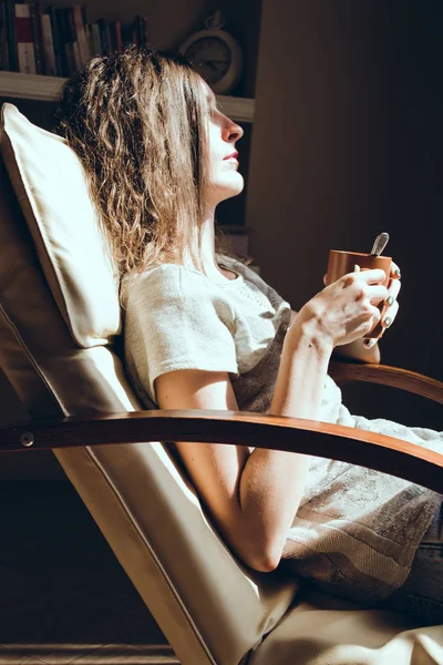 Evde zaman zevk. Kadın portre rahatlatıcı pencere holding bir fincan çay veya kahve yakın konforlu modern sandalyede kapatın. Doğal ışık. Rahat ev. Anın tadını çıkar. Parlak güneşli bir günde. Fincan sıcak içecek