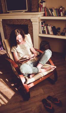 Evde zaman zevk. Kadın kağıt kitap okuma konforlu modern sandalye rahatlatıcı. Doğal ışık. Rahat ev. Anın tadını çıkar. Parlak güneşli bir günde. Bilgi, öğrenme kavramı. Kitap kurdu öğrenci. Dikey