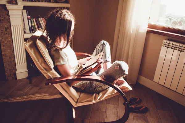 Evde zaman zevk. Kağıt kitap okuma penceresi yakınındaki rahat modern sandalye rahatlatıcı bir kadın. Doğal ışık. Rahat ev. Anın tadını çıkar. Parlak güneşli bir günde. Bilgi ve kavram öğrenme. Kitap kurdu öğrenci