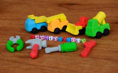 Ahşap doku arka plan üzerinde sentetik kauçuk yapılmış inşaat ağır makine ve inşaat araçları, minyatür modeli. Ortadaki küçük plastik blok üzerine basılmış inşaat metni.  