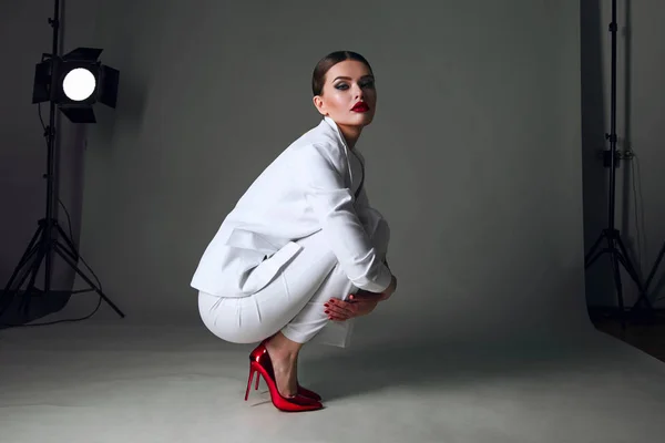 Beautiful young girl with a beautiful figure in a white trouser suit and red shoes sits in a beautiful pose on the floor in the studio.