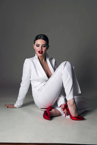 Beautiful young girl with a beautiful figure in a white trouser suit and red shoes sits in a beautiful pose on the floor in the studio.