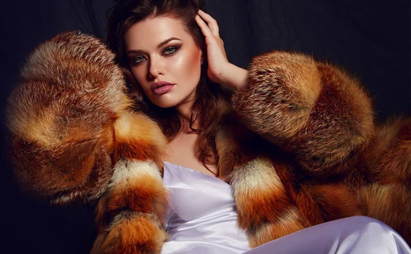 A young glamorous woman with long brown hair and a white satin slip dress in luxurious fluffy furs. Glamorous life. Advertising of fur products.