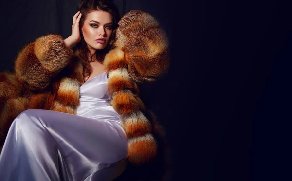 A young glamorous woman with long brown hair and a white satin slip dress in luxurious fluffy furs. Glamorous life. Advertising of fur products.