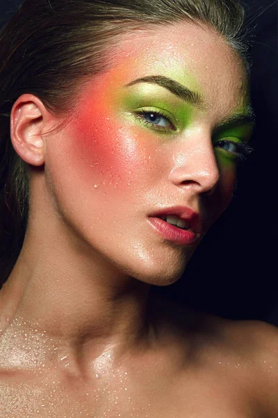 Beautiful young attractive girl lies. A large portrait is a face. Bright colorful make-up - green and pink colors. Drops of water on the skin, splashes, wet face. Cosmetics, healthy skin.