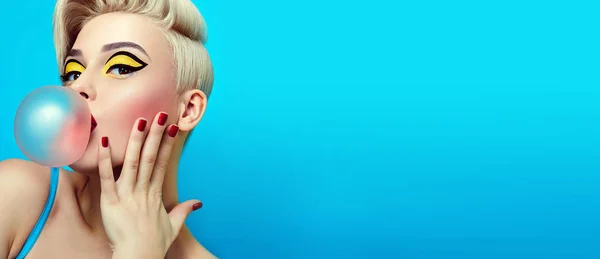 Fashionable girl with a stylish haircut inflates a chewing gum. The girl in the studio on a blue background. The girl's face with bright makeup and yellow with black shadows on the eyes.