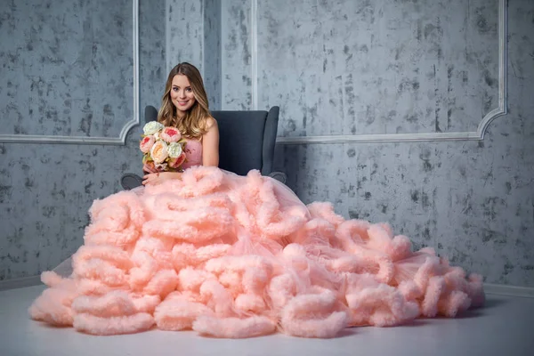 Hermosa joven con el pelo ondulado largo sosteniendo un ramo de flores en rosa vestido de la nube de observación está sentado en la silla en el interior — Foto de Stock