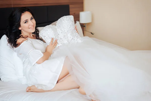 Nevěsta sedí na posteli a v rukou drží svatební šaty — Stock fotografie
