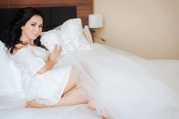 La novia se sienta en la cama y mantiene el vestido de novia en las manos — Foto de Stock