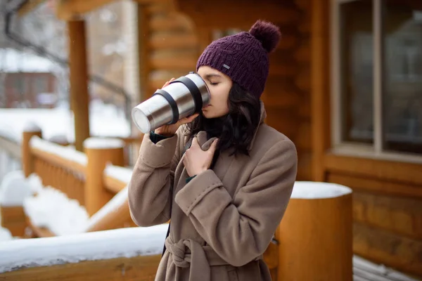 Una chica sosteniendo un termo con una bebida caliente. Una taza de café en sus manos. Callejón de invierno en la ciudad, una joven camina por la ciudad —  Fotos de Stock