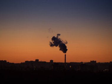 Endüstriyel bitki ve şehir siluetinin dumanlı dumanlı tek gişe bacası. Arka planda açık günbatımı gökyüzü var.