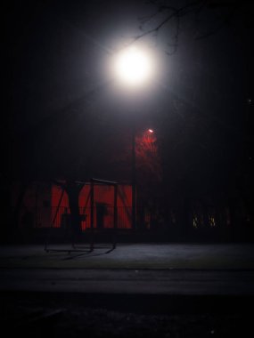 Gece vakti tek bir sokak lambasıyla aydınlatılan futbol gollü boş okul parkı. Çitli karanlık avlu ve karanlıkta yalnız sokak lambası. Uzun pozlu çekim.