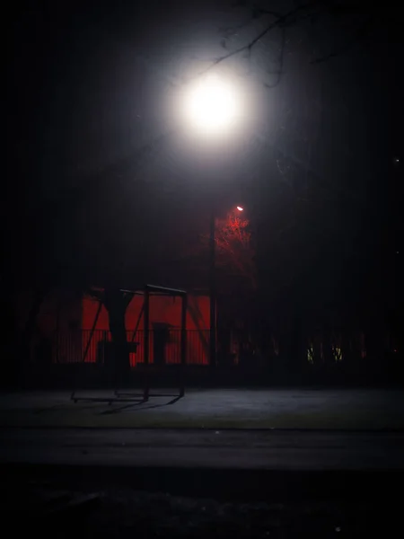 Gece vakti tek bir sokak lambasıyla aydınlatılan futbol gollü boş okul parkı. Çitli karanlık avlu ve karanlıkta yalnız sokak lambası. Uzun pozlu çekim.