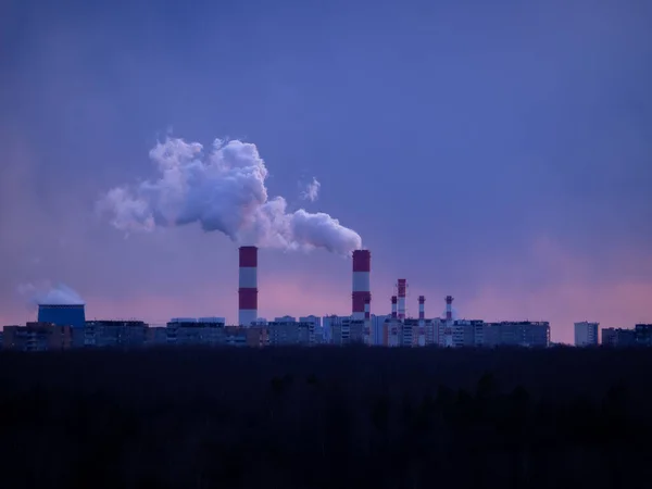 Cityscape silhouette with clouds of smoke from smoking chimneys of thermal power station and pipes of the industrial plant and clear blue sky at sunset