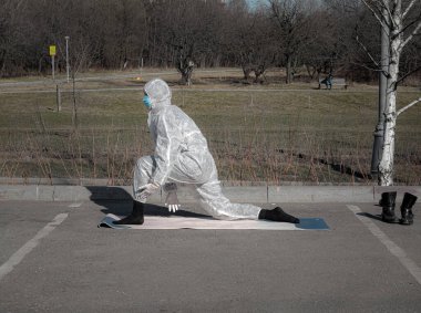 Salgın sırasında hayat. Koruyucu giysili, maskeli ve eldivenli bir kadın boş şehir parkında yoga egzersizleri yapıyor. Yaz sezonu, seçici odaklanma. Coronavirus (covid-19) salgını sırasında yaşam tarzı ve spor.