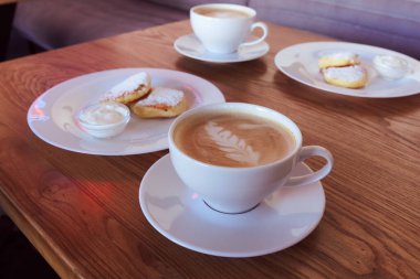 Ahşap zemin üzerinde latte sanat ve peynir krepli iki fincan kapuçino. Güzel köpük, beyaz bardaklar, metin için yer.