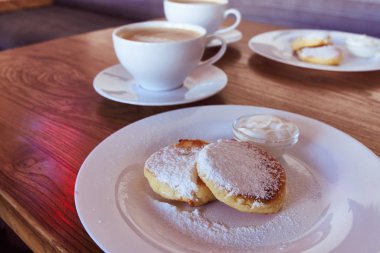 Gurme kahvaltısı, süzme peynirli krep, şurup. Beyaz tabak ve kapuçino ile pudra şekerli Ukrayna yemeği