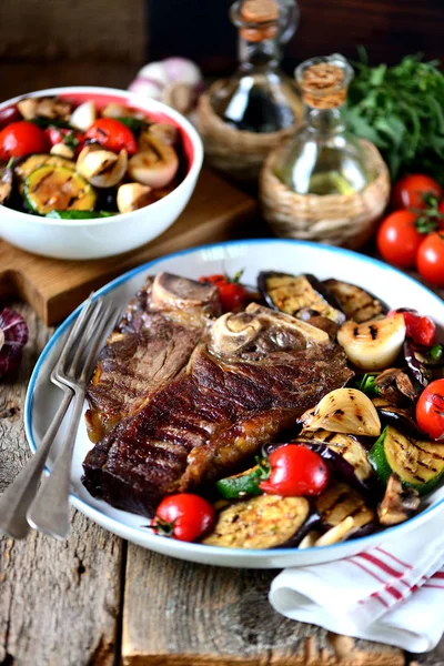 Grilled t-bone steak with grilled vegetables on an old wooden background. Rustic style. — Stock Photo, Image
