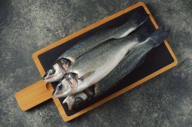 Gri arka planda servis panosunda üç taze çiğ levrek. Üst görünüm. 