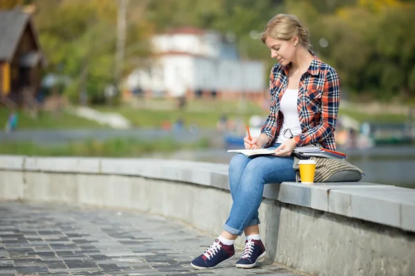 Portrait of an attractive student girl sitting at bridge writing — Stock Photo, Image