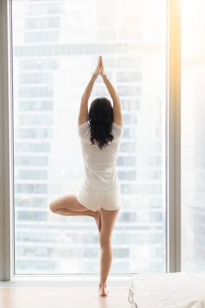 Young attractive woman in Vrksasana pose near the floor window